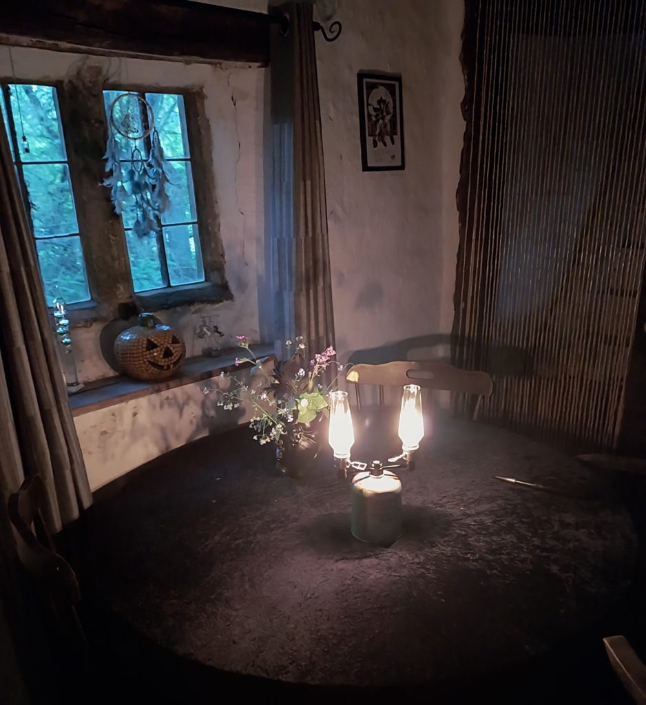 A picture of a double burner lamp, attached to a camping gas cannister. It sits on a round table with a black velvet tablecloth, in front of an old cottage window. On the windowsill sits a mosaic pumpkin ornament with a dreamcatcher hanging above it.