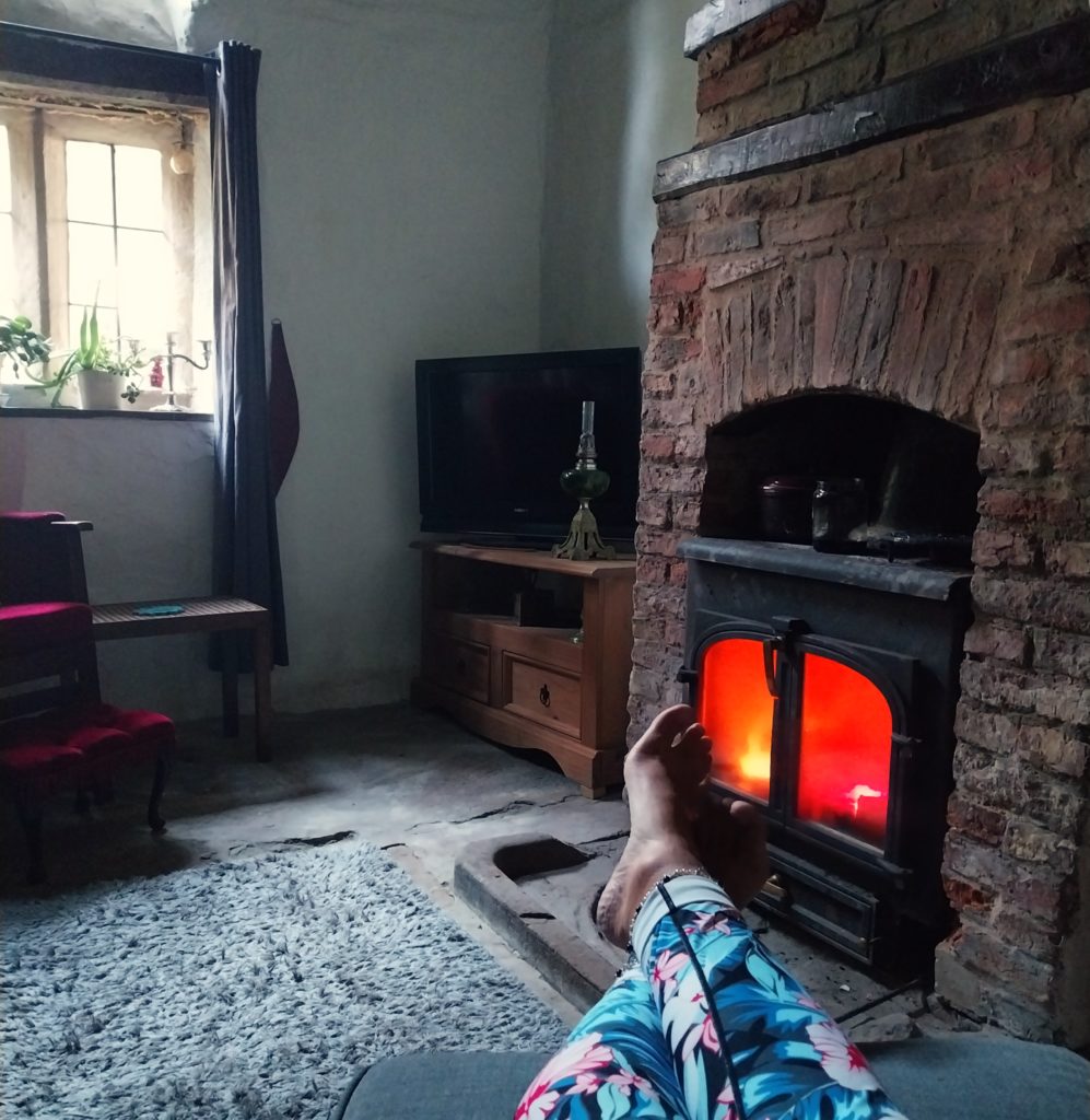 A glowing woodburner in a cosy cottage, with my feet sticking out on a tuffet in front of it.