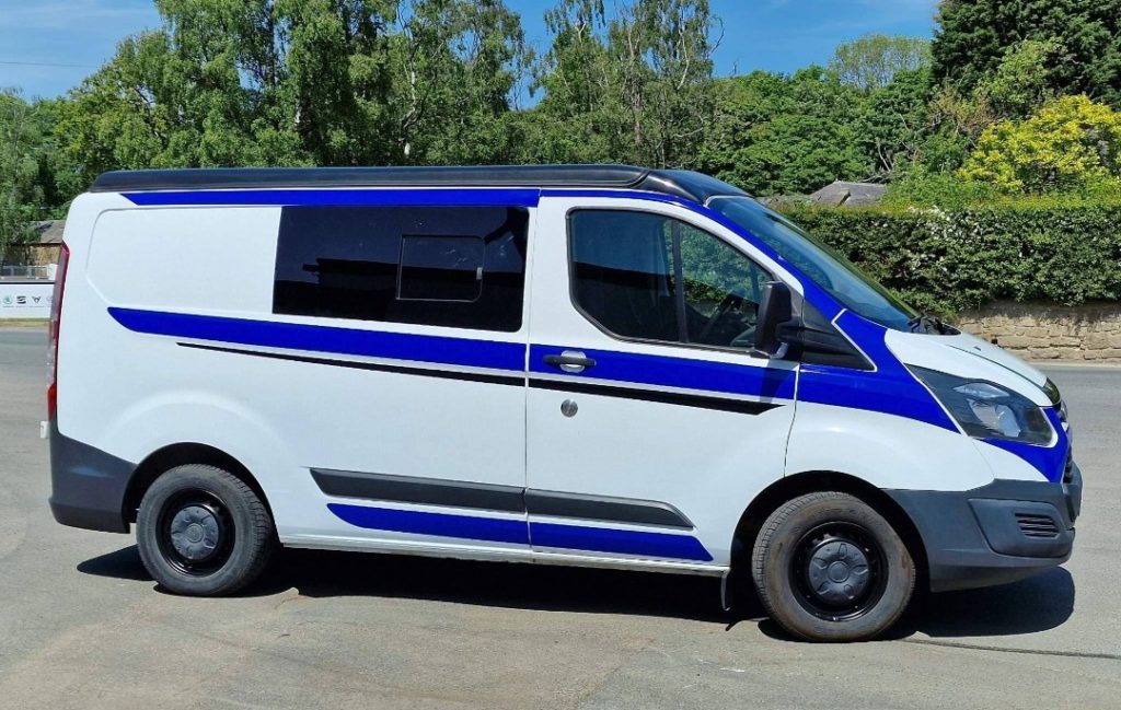 A side view of a white ford transit custom van with blue racer stripes.
